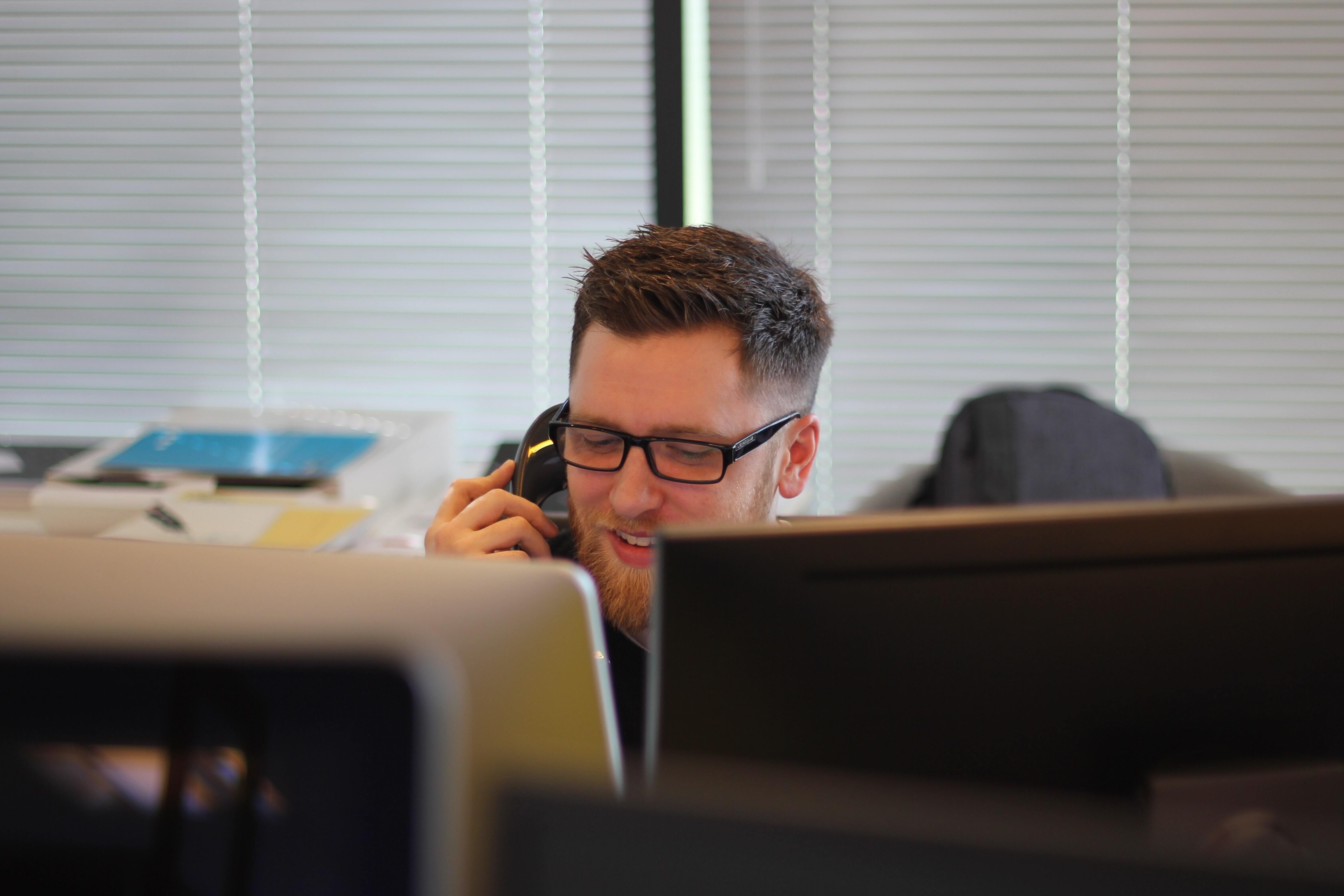 Man on the Phone Smiling While Looking at Computer Screen in the Office