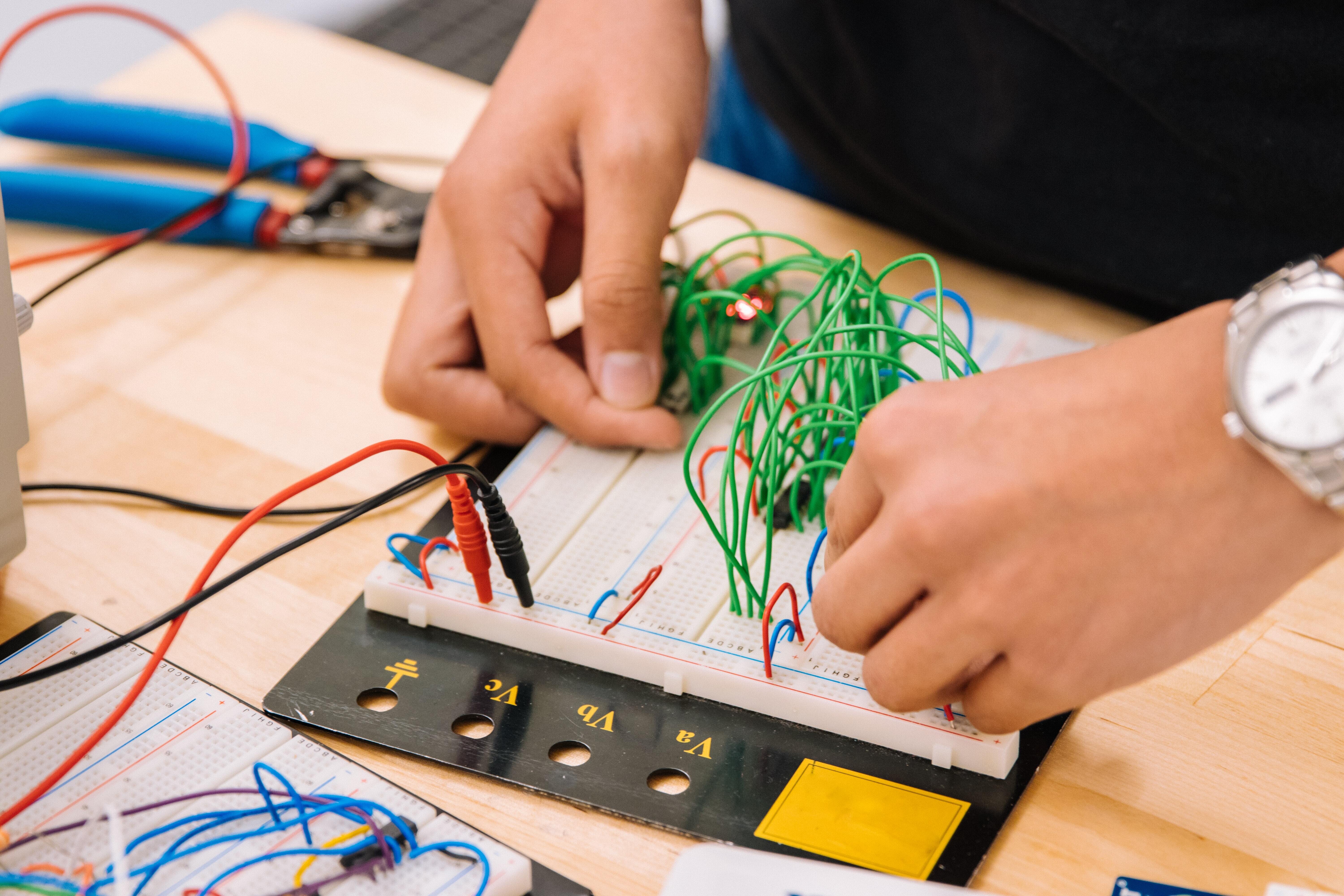 Two Hands Connecting Wires to a Circuit Board Placed on a Wooden Table