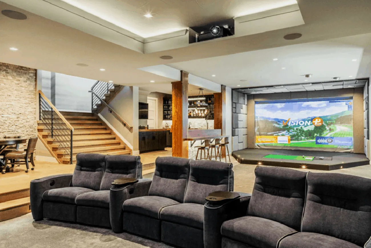 A basement golf room with a stacked stone wall, wood stairs, and recliners facing the simulator