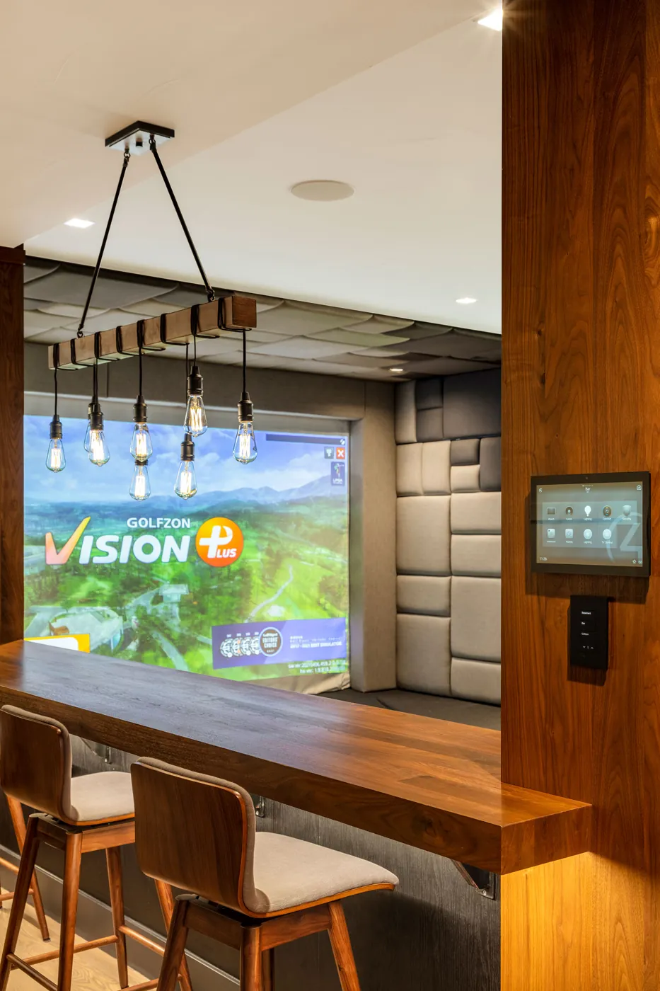 A walnut bar with stools and pendant lights beside a padded golf simulator bay